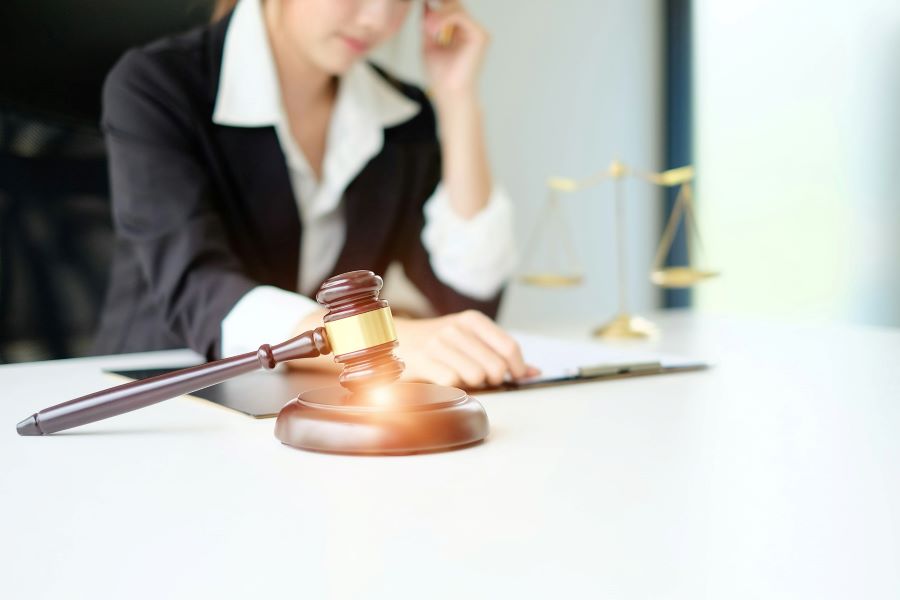 a woman in a suit sitting with a gavel in the foreground and scales of justice in the background